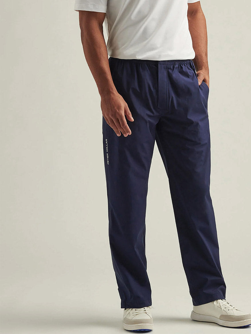 A man wearing a white short-sleeve shirt, Peter Millar Shield Pull-On Rain Pant in navy blue, and white sneakers stands against a plain background.