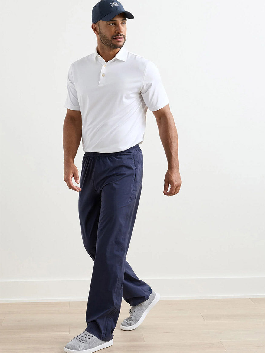 A man wearing a white polo, Peter Millar Shield Pull-On Rain Pant in navy, gray sneakers, and a navy cap walks indoors on a light wood floor.
