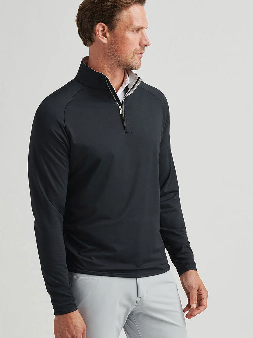 Man wearing a Peter Millar Stealth Performance Quarter-Zip in black and light gray pants, standing and looking to the side against a plain background.