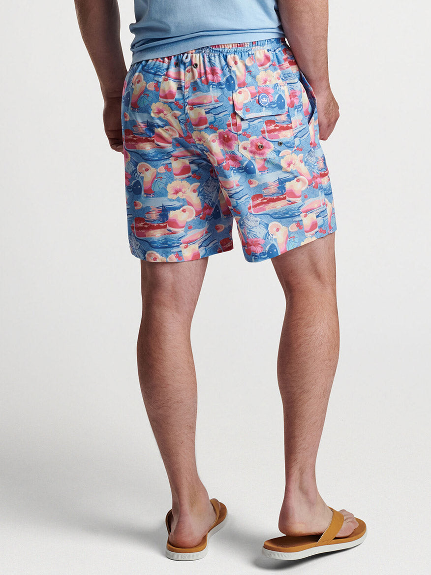 A man wearing exclusive Peter Millar Tequila Sunrise Swim Trunks in Infinity with patterned prints and flip-flops, viewed from behind.