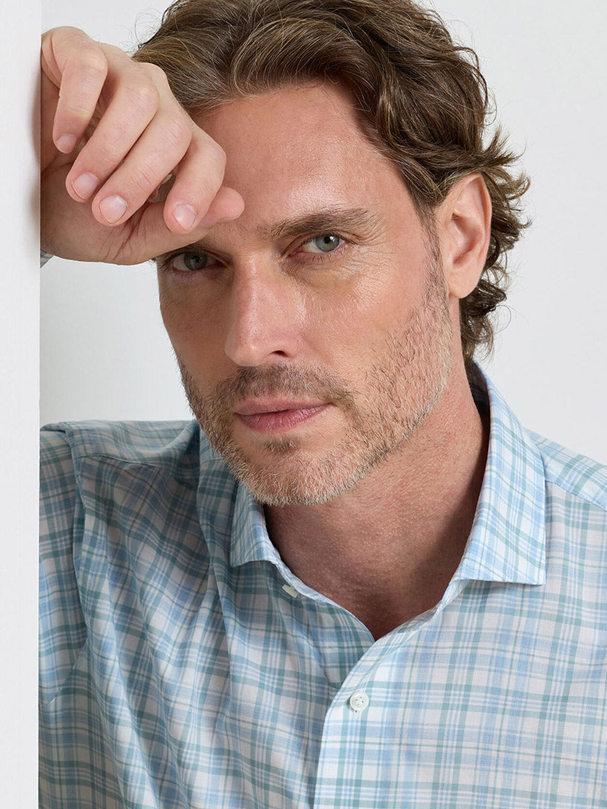 A man with light brown hair and a trimmed beard wears the Peter Millar Tropea Italian Poplin Sport Shirt in Light Willow, resting his hand on his forehead and looking at the camera against a plain background.