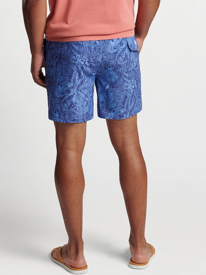 A man in Peter Millar Tropicrazy Swim Trunk in Atlantic Blue and flip-flops standing with his hands in his pockets.
