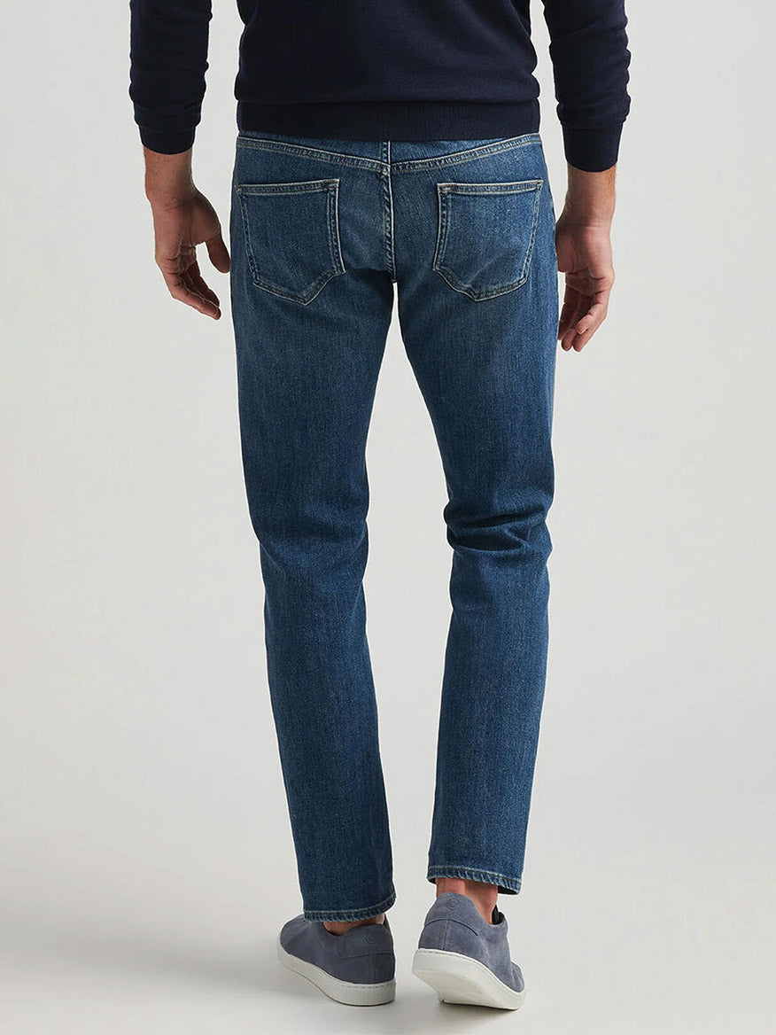 A person wearing Peter Millar Vintage Washed Five-Pocket Denim in True Blue Wash, a dark sweater, and gray slip-on shoes stands with their back to the camera against a plain background.