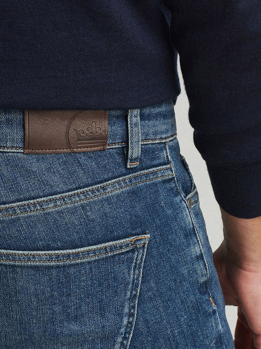 A person wearing Peter Millar Vintage Washed Five-Pocket Denim in True Blue Wash, featuring an Italian leather back patch by the waistband, paired with a dark long-sleeve shirt.