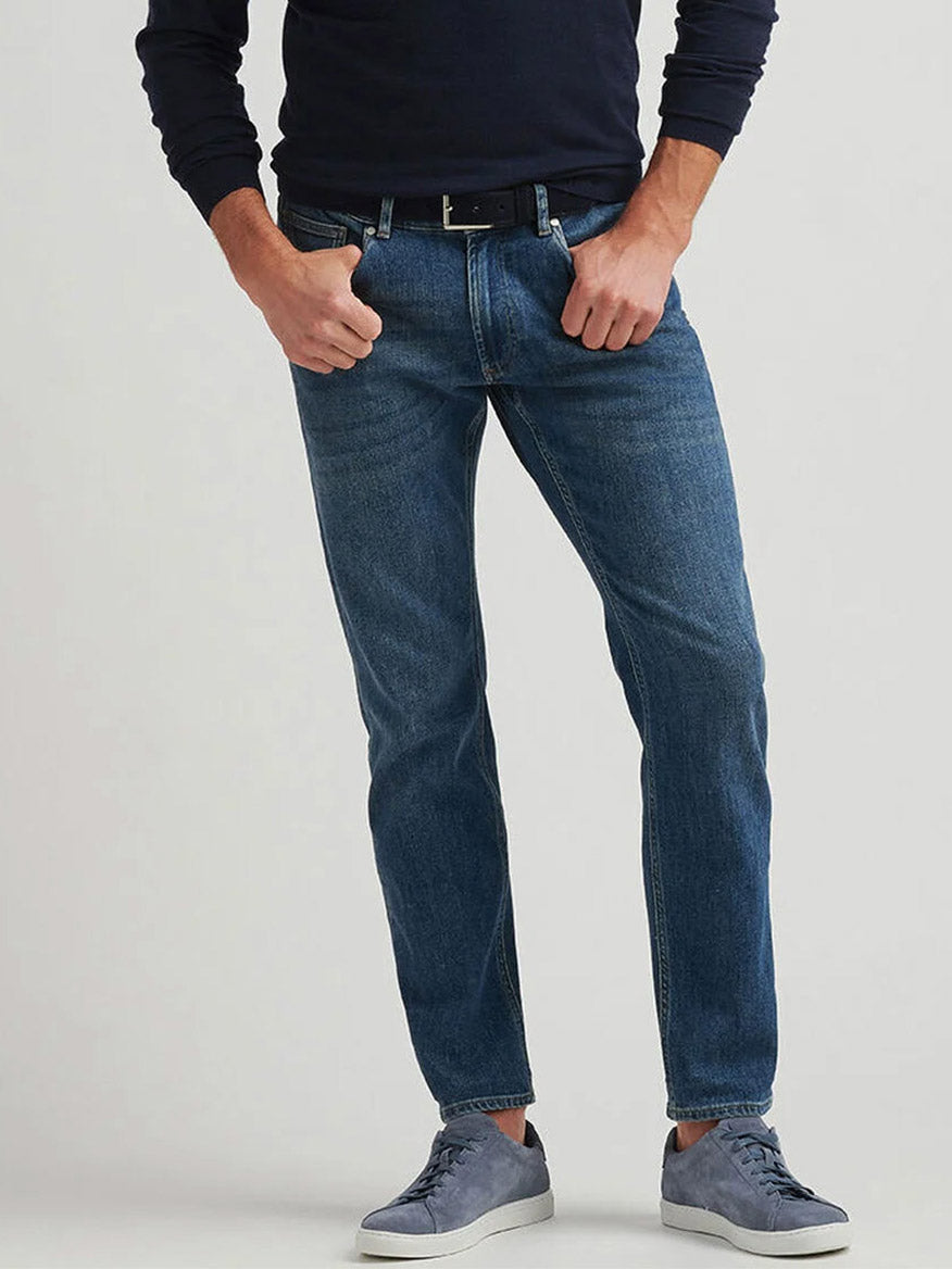 A person wearing a black long-sleeve shirt, a black belt, gray sneakers, and Peter Millar Vintage Washed Five-Pocket Denim in True Blue Wash stands against a light background.