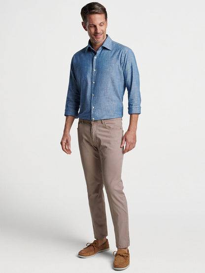 A man smiling, standing in a studio, wearing a blue button-up shirt made of lightweight fabric, Peter Millar Wayfare Five-Pocket Pant in Khaki, and brown shoes.