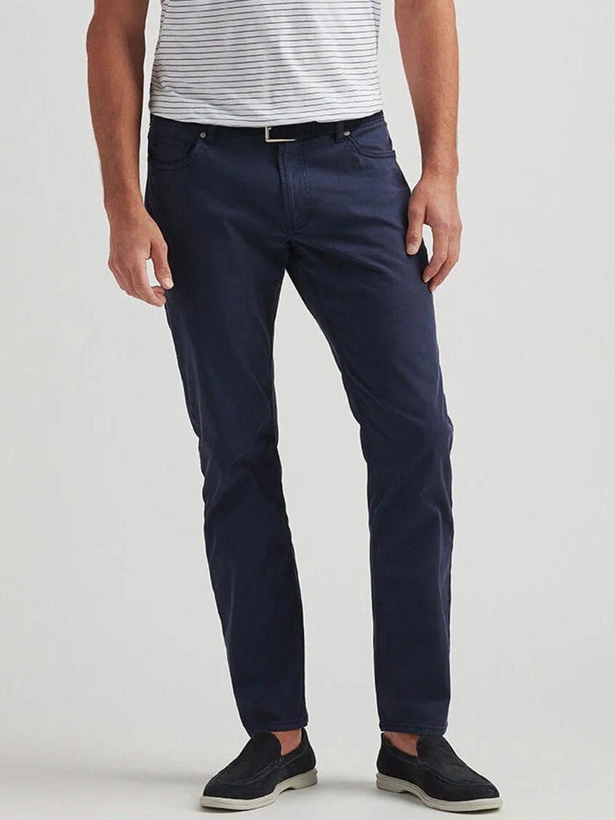 A person wearing a short-sleeve striped shirt, Peter Millar Wayfare Five-Pocket Pant in Navy, black slip-on shoes, and a black belt stands against a plain background.