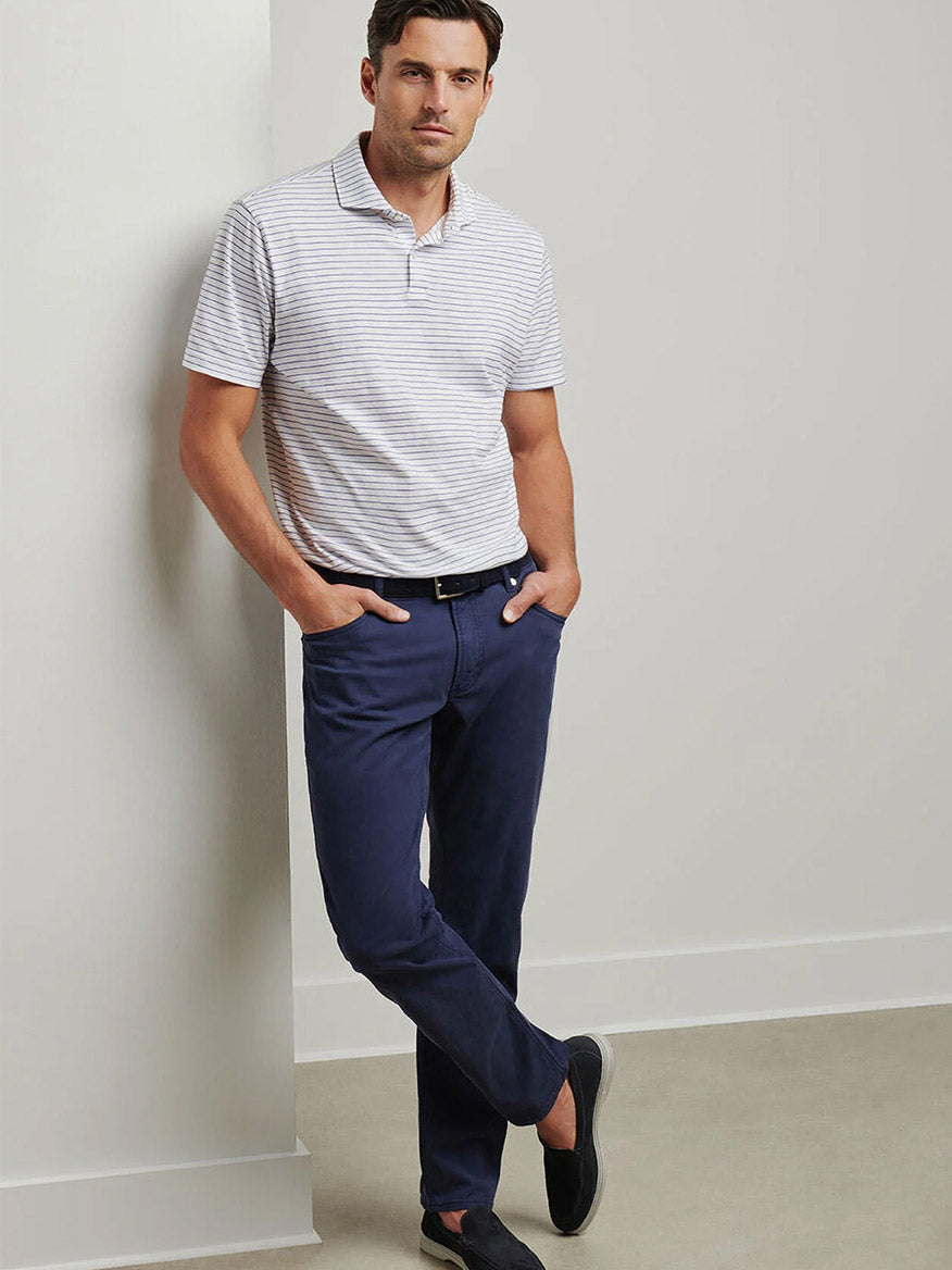 A man stands against a wall wearing a short-sleeve striped polo, the Peter Millar Wayfare Five-Pocket Pant in Navy (noted for wrinkle resistance), a black belt, and dark loafers, hands in his pockets.