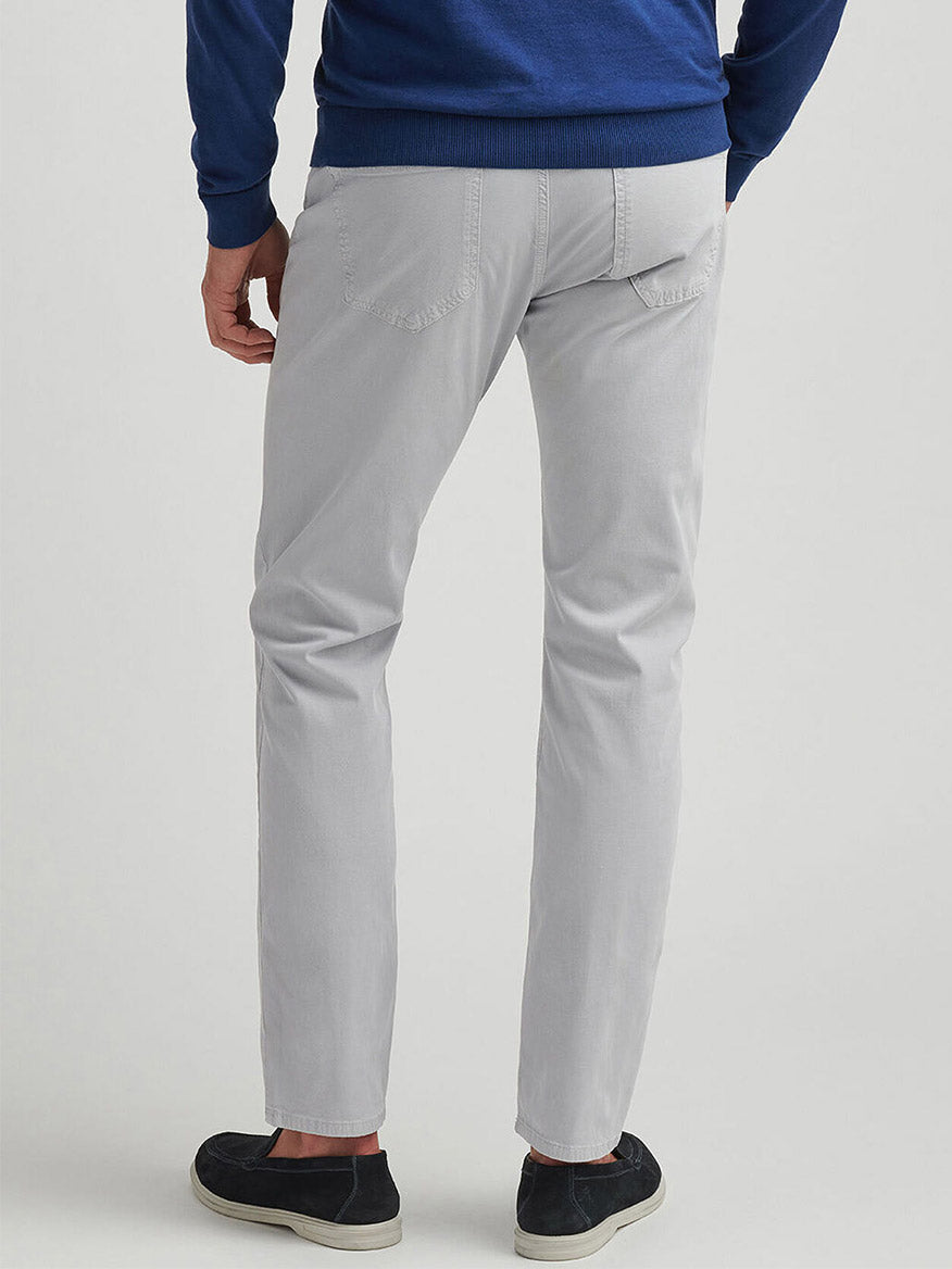 A person stands facing away, wearing Peter Millar Wayfare Five-Pocket Pant in Platinum, a blue sweater, and black slip-on shoes against a plain background.