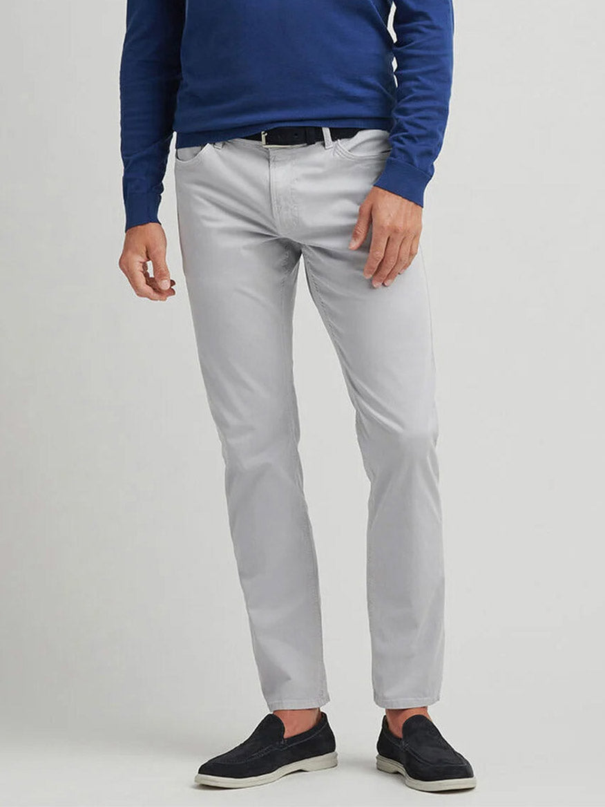 A person wearing a blue long-sleeve shirt and Peter Millar Wayfare Five-Pocket Pant in Platinum with black slip-on shoes stands against a plain background.