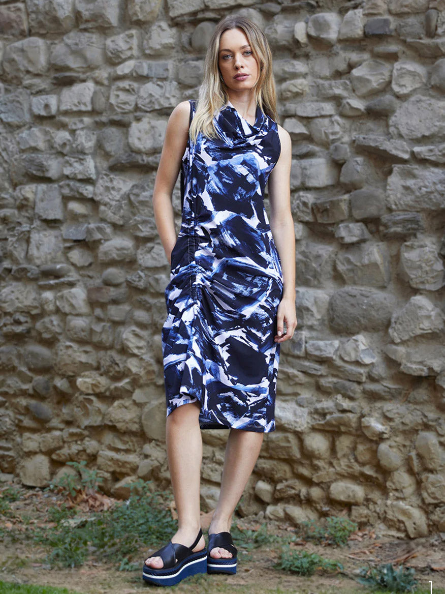 A woman wearing the Porto Indira Ruched Dress in Shadow Shodo and black platform sandals stands in front of a stone wall.
