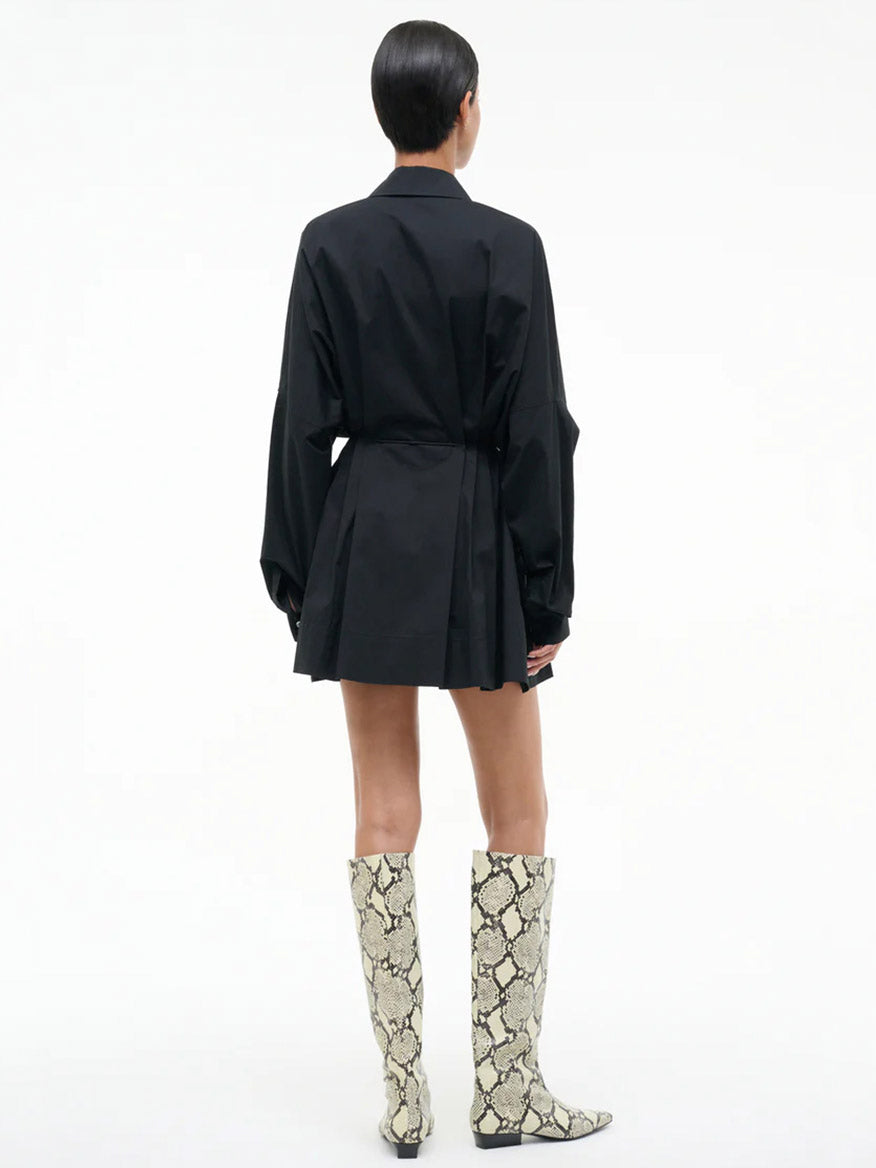 A person stands with their back to the camera wearing the STAUD Chi Dress in Black, featuring an adjustable waist tie, paired with knee-high white snake-print boots against a plain white background.