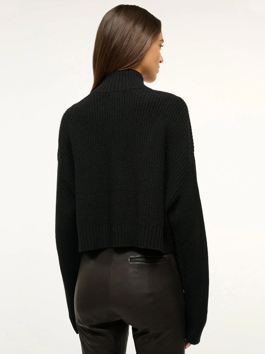 Woman with straight brown hair wears the STAUD Cropped Hampton Cashmere Sweater in Black, a long-sleeve, chunky knit style, paired with black leather pants, shown from the back against a plain white background.