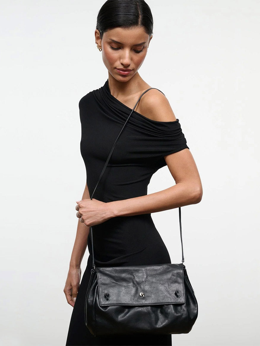 A person in a black off-the-shoulder dress wearing a black STAUD Cy Convertible Bag, a leather shoulder bag with a fold-over flap that can be worn as a crossbody.