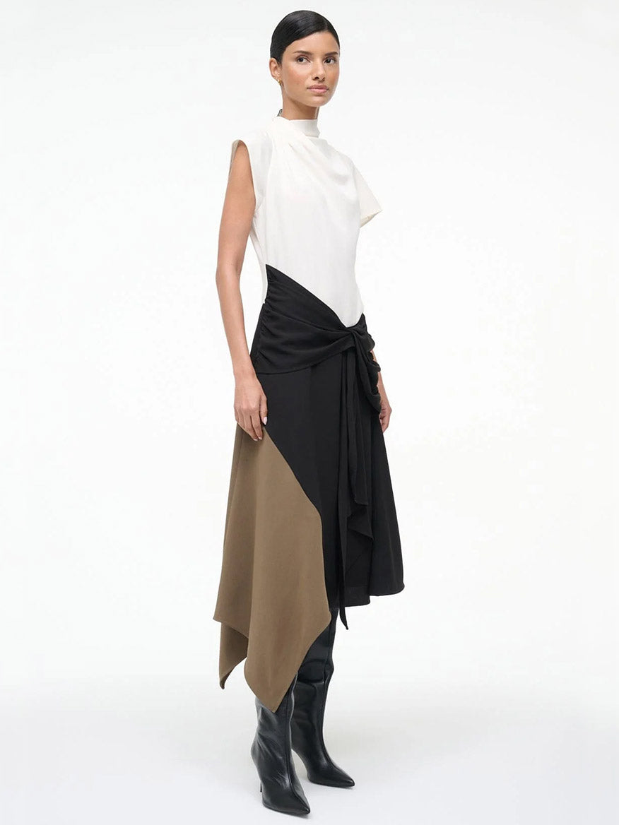 A woman wears the STAUD Davina Dress in Black Multi—a sleeveless, high-neck, asymmetrical dress with a handkerchief hem—styled with black knee-high boots, posed against a plain background.