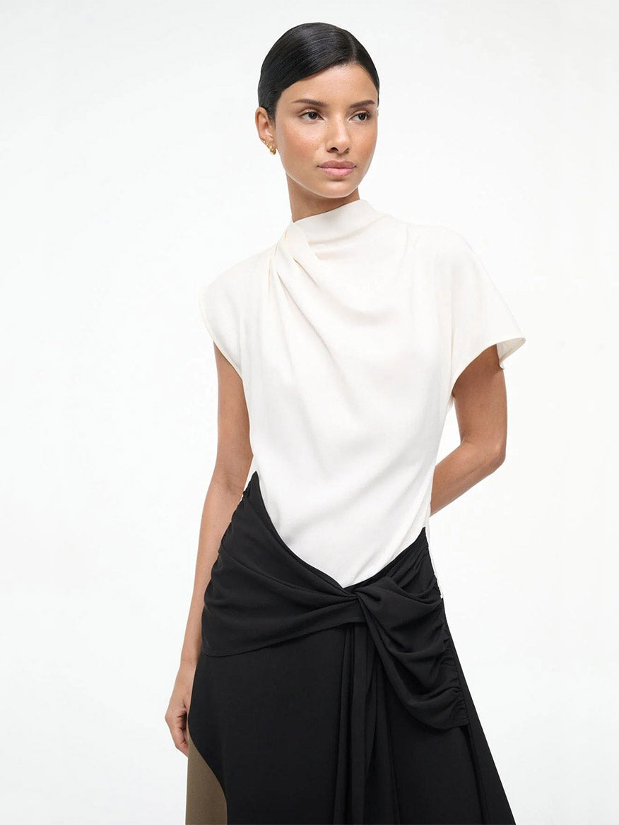 A woman stands against a plain background wearing the STAUD Davina Dress in Black Multi, which features a white short-sleeve top with a high neckline and a black asymmetrical skirt with a draped knot detail at the waist.