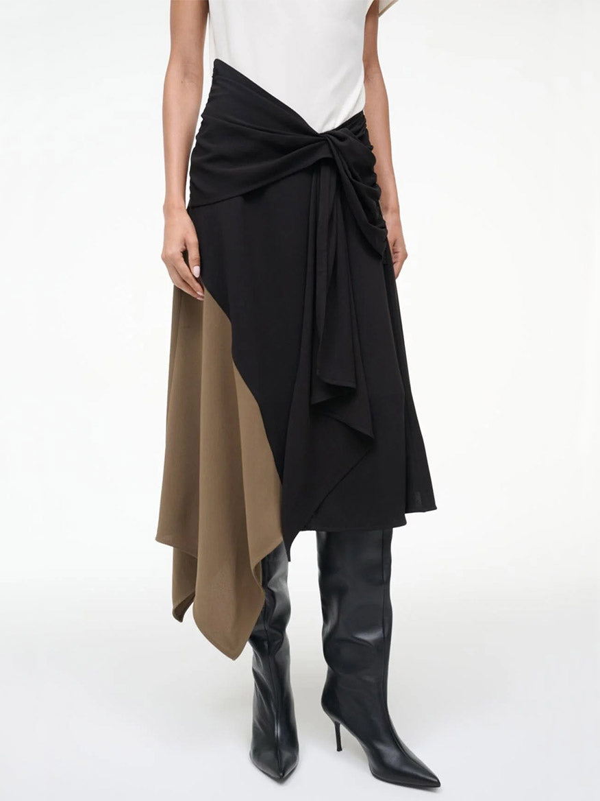 A woman wears the STAUD Davina Dress in Black Multi, featuring a black and brown asymmetrical handkerchief hem and knot detail, paired with black knee-high boots.