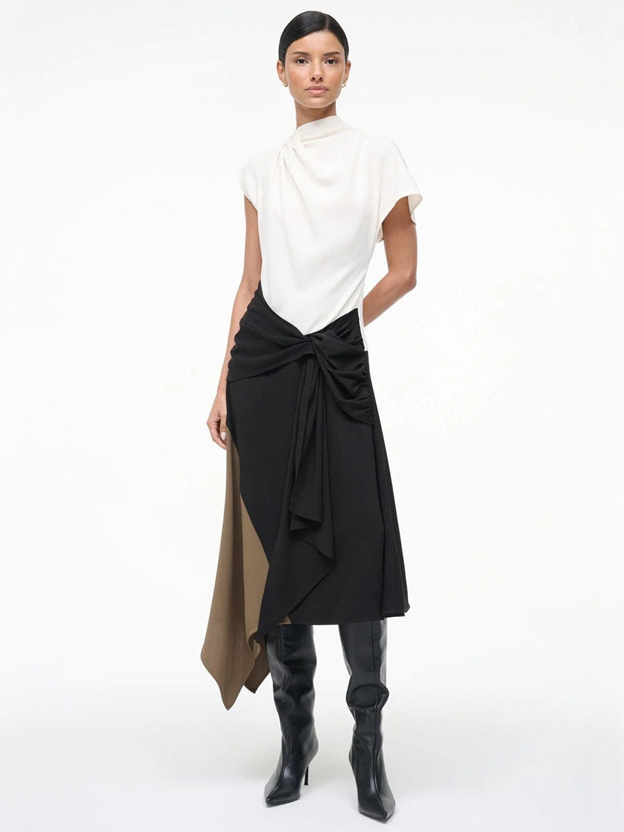 A woman stands against a plain background wearing the STAUD Davina Dress in Black Multi, featuring a high-neck top and color-blocked handkerchief hem, paired with black knee-high boots.