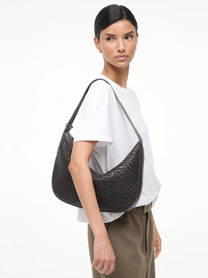 A woman in a white t-shirt and beige pants carries the STAUD Large Demy Shoulder Bag in Espresso, featuring an adjustable shoulder strap, while standing against a plain white background.