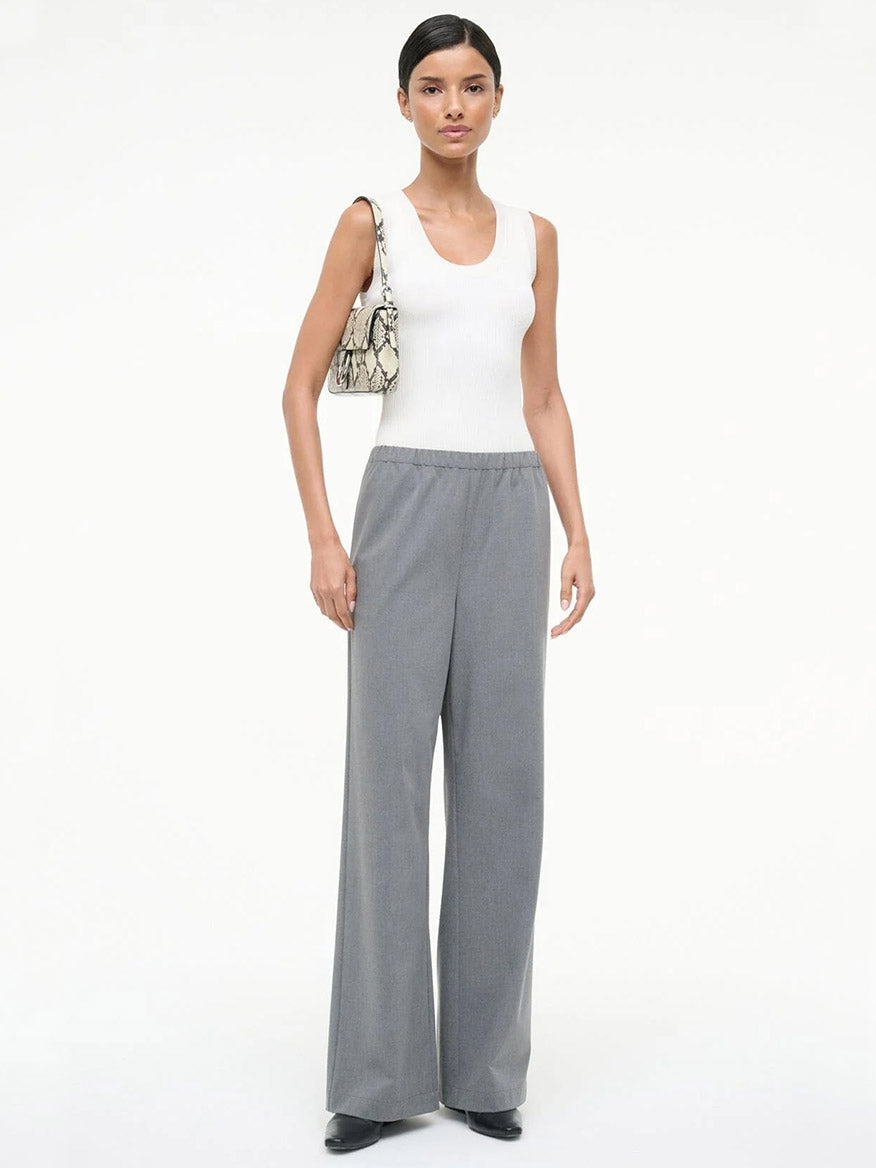 A woman stands in a white sleeveless top, STAUD Denali Pant in Static, black shoes, and carries a snakeskin-pattern shoulder bag against a plain white background.