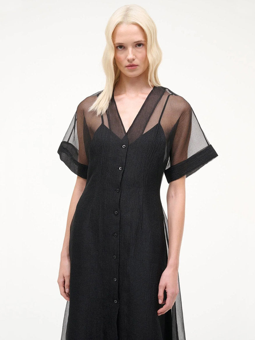 A woman wearing the STAUD Elle Dress in Black, a sheer organza midi dress with short sleeves, stands against a plain light background.