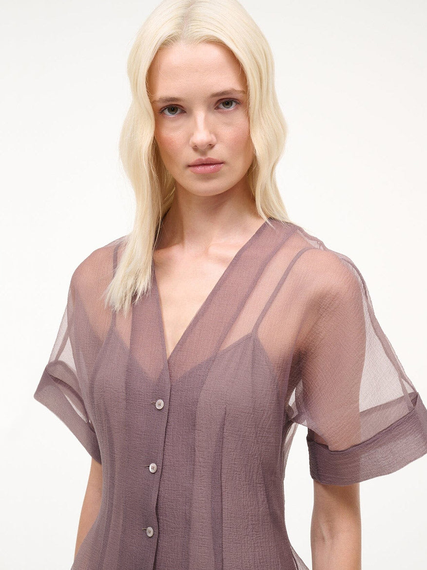 A woman with long, straight blonde hair wears a sheer short-sleeved STAUD Elle Dress in Mink—a V-neck A-line style with front buttons—standing against a plain light background.