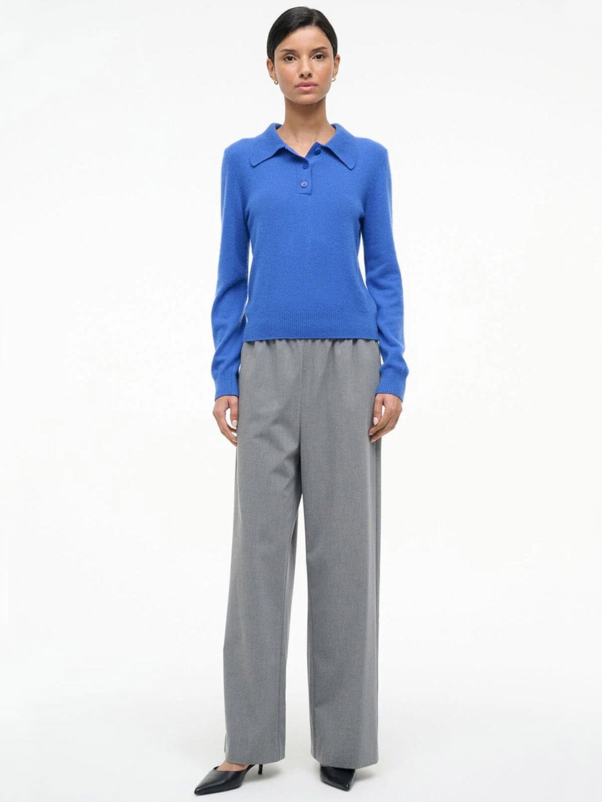 A woman faces forward, wearing a STAUD Gemma Cashmere Sweater in Sapphire, gray wide-leg trousers, and black pointed shoes against a plain white background.