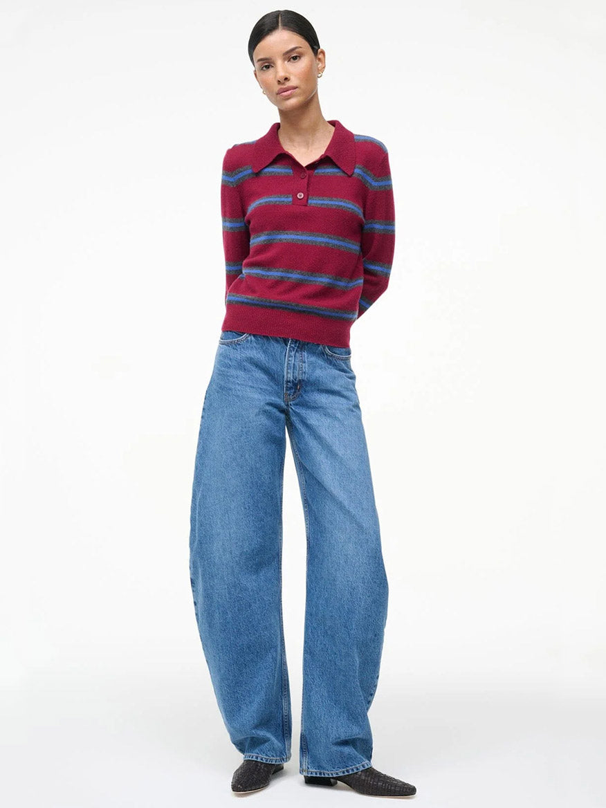 Woman standing against a white background, wearing a STAUD Gemma Cashmere Sweater in Crimson Stripe, wide-leg blue jeans, and dark shoes.