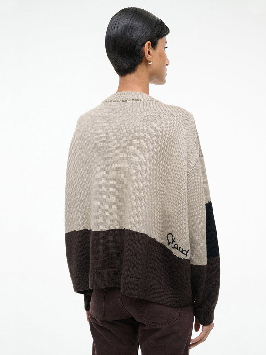 A person with short dark hair, wearing the STAUD Giada Sweater in Stables—a loose beige and brown color-block crewneck with "Staud" embroidered near the hem—stands facing away from the camera.