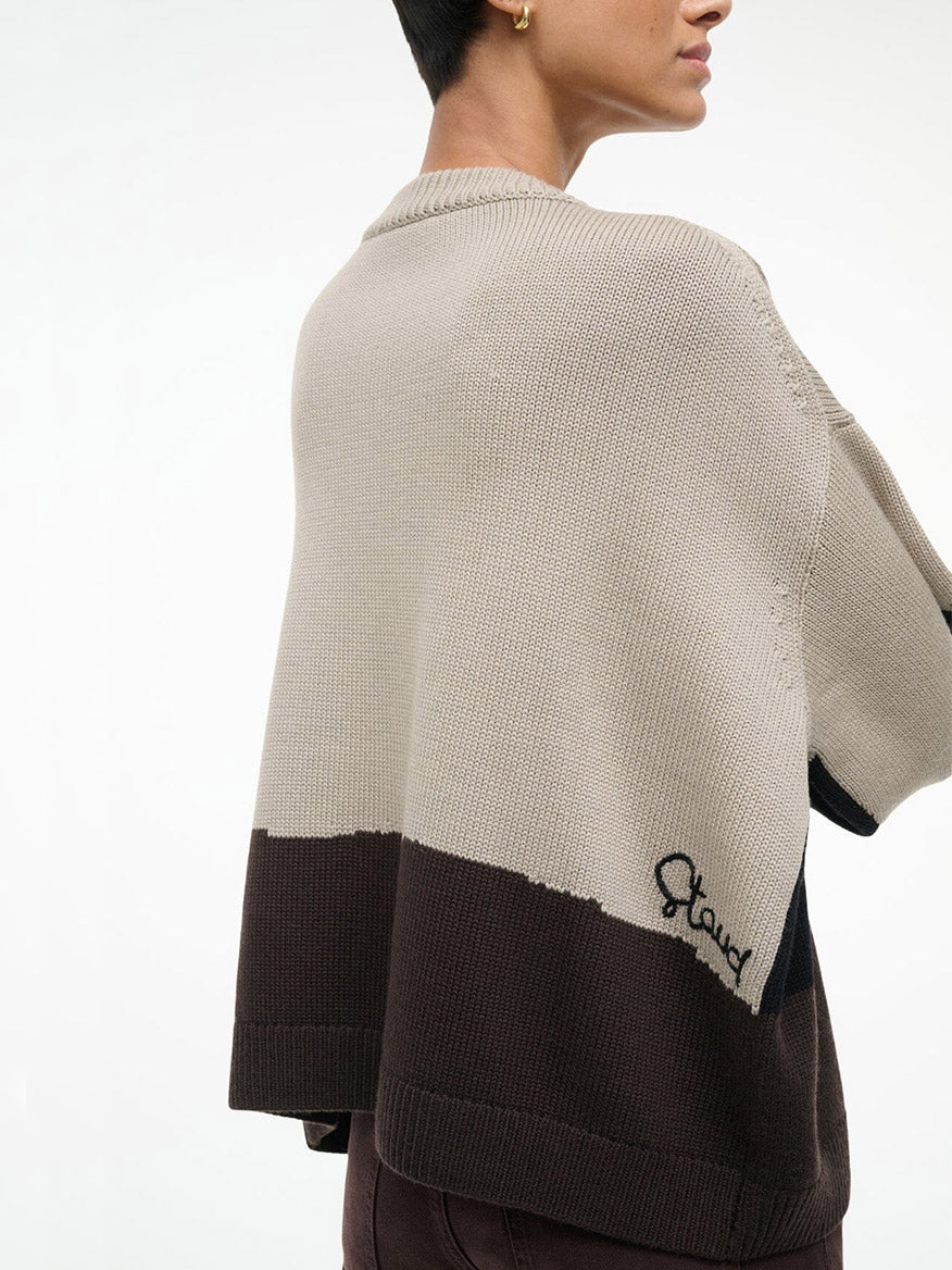 Person wearing the STAUD Giada Sweater in Stables, a color-blocked beige and brown crewneck with "Stand" embroidered near the hem, shown from the back and side. Made from soft merino wool for comfort and style.