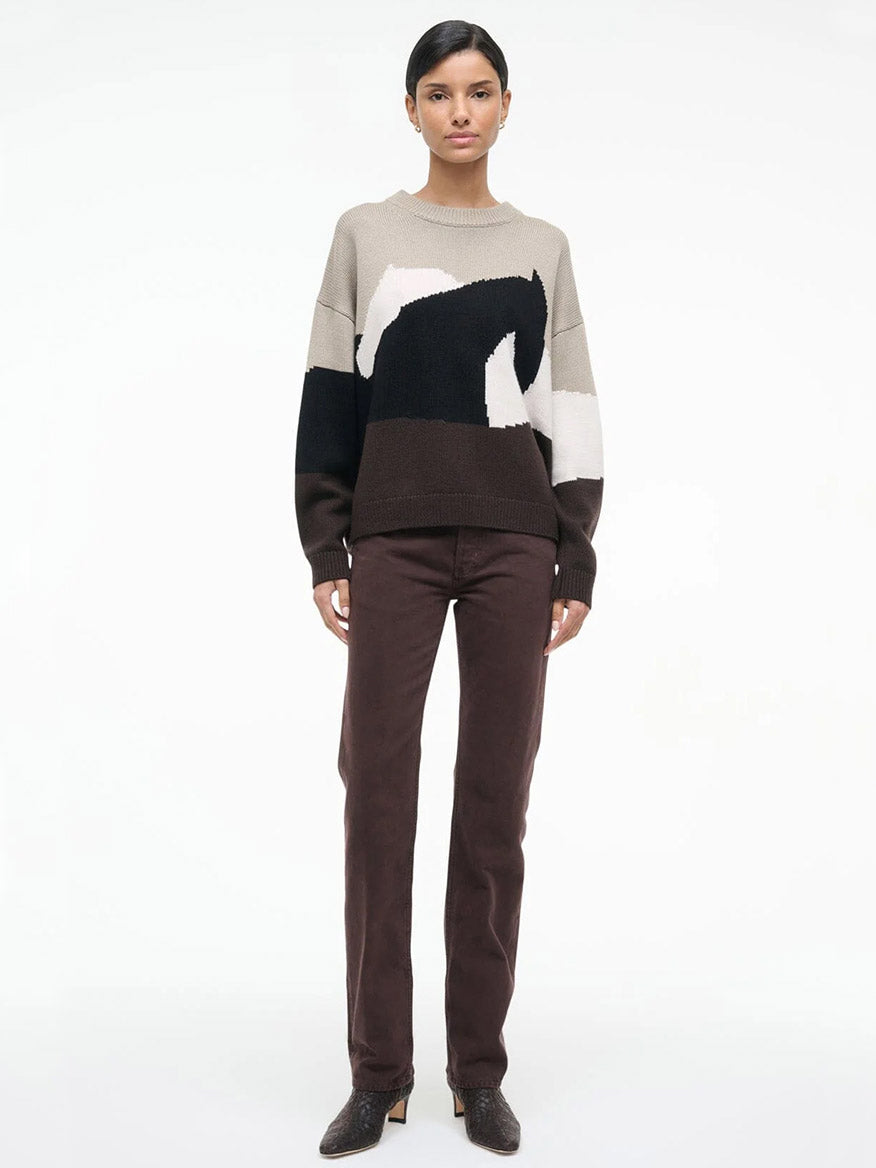 A woman stands against a plain background wearing the STAUD Giada Sweater in Stables, featuring a beige, black, and white abstract horse pattern, paired with dark brown pants and heeled shoes.