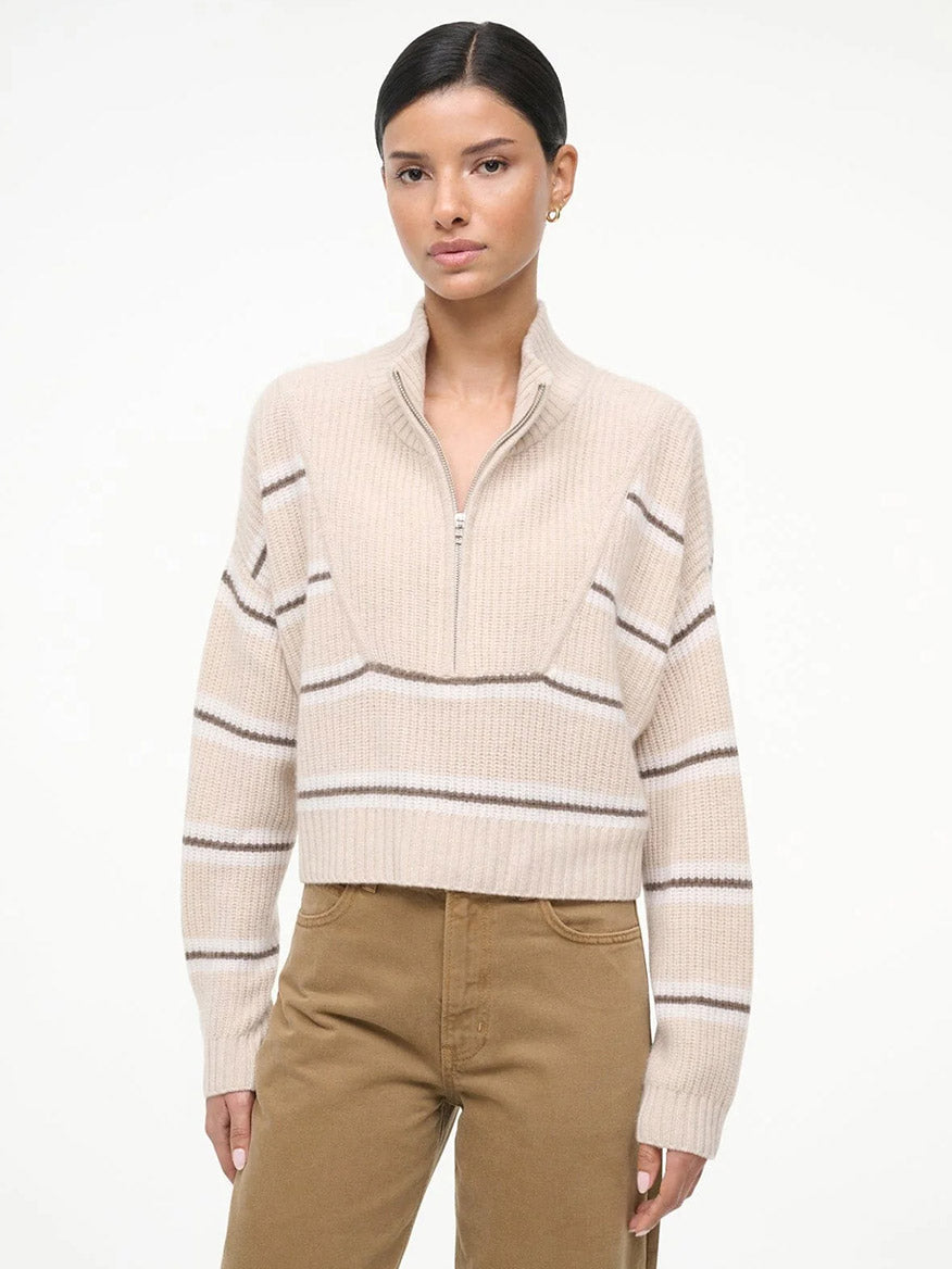 A woman stands against a white background wearing the STAUD Hampton Cropped Cashmere Sweater in Whisper Stripe with beige stripes and brown pants, looking directly at the camera with a neutral expression.