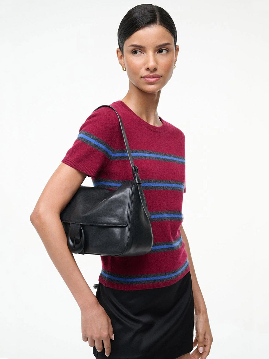 A woman wears a short-sleeve red and blue striped sweater, black skirt, and carries the STAUD Harlow Medium Bag in Black with a Moon buckle, standing against a plain white background.