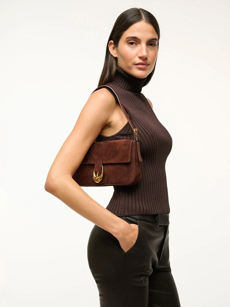 Woman standing sideways in a sleeveless brown turtleneck top, black pants, and carrying the STAUD Harlow Bag in Mahogany Suede, featuring a small shoulder design and gold moon buckle detail.
