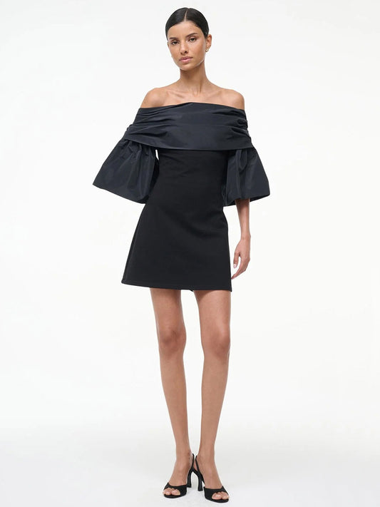 A woman stands against a plain white background wearing the STAUD Harper Dress in black, an off-the-shoulder ponte mini dress with voluminous taffeta sleeves, styled with black heels.