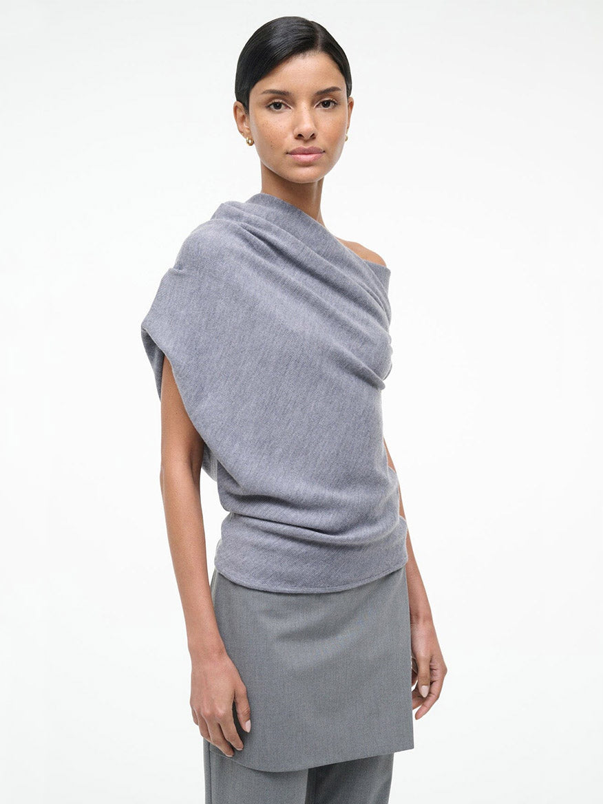 A woman stands against a plain background wearing the STAUD Mabel Sweater in Heather Grey, styled off one shoulder, paired with a matching grey skirt.