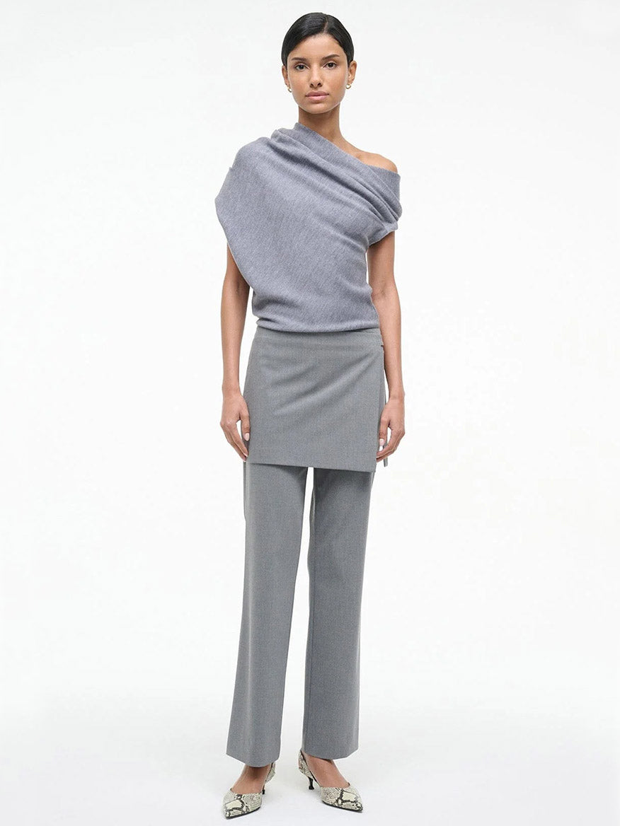 A woman stands against a white background wearing the STAUD Mabel Sweater in Heather Grey, paired with a matching grey skirt layered over trousers and snakeskin-patterned heels.