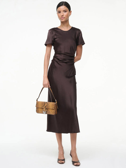 A woman wearing the STAUD Marcel Silk Dress in Earth and black heels holds a tan snakeskin handbag, standing against a white background.