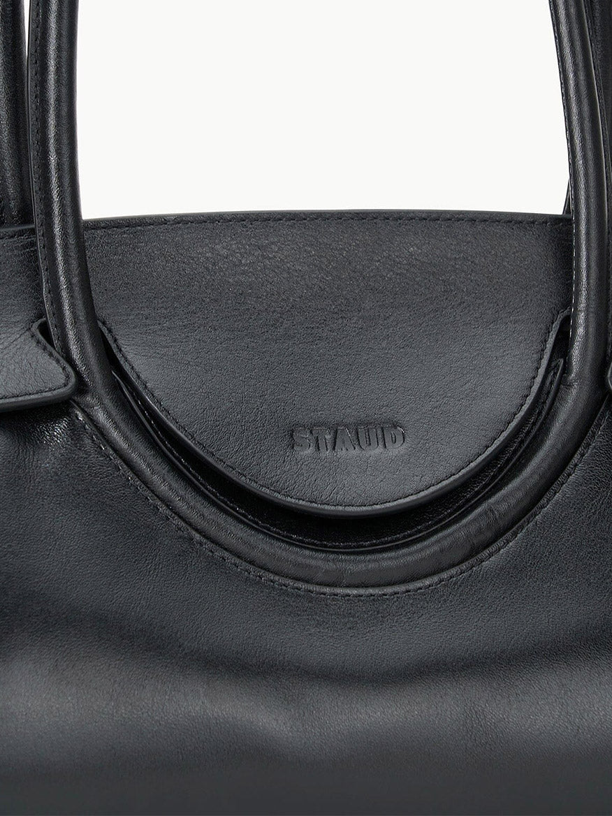 Close-up of the STAUD Maude Shoulder Bag in Black, crafted from polished leather with rounded handles, an east-west silhouette, and "STAUD" embossed on the front flap.