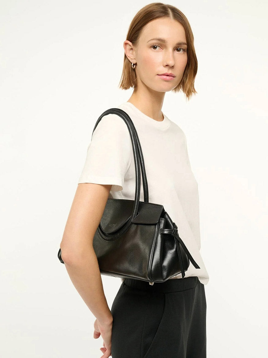 A woman with straight, shoulder-length hair wears a white t-shirt and black pants, holding the STAUD Maude Shoulder Bag in Black against a plain background.