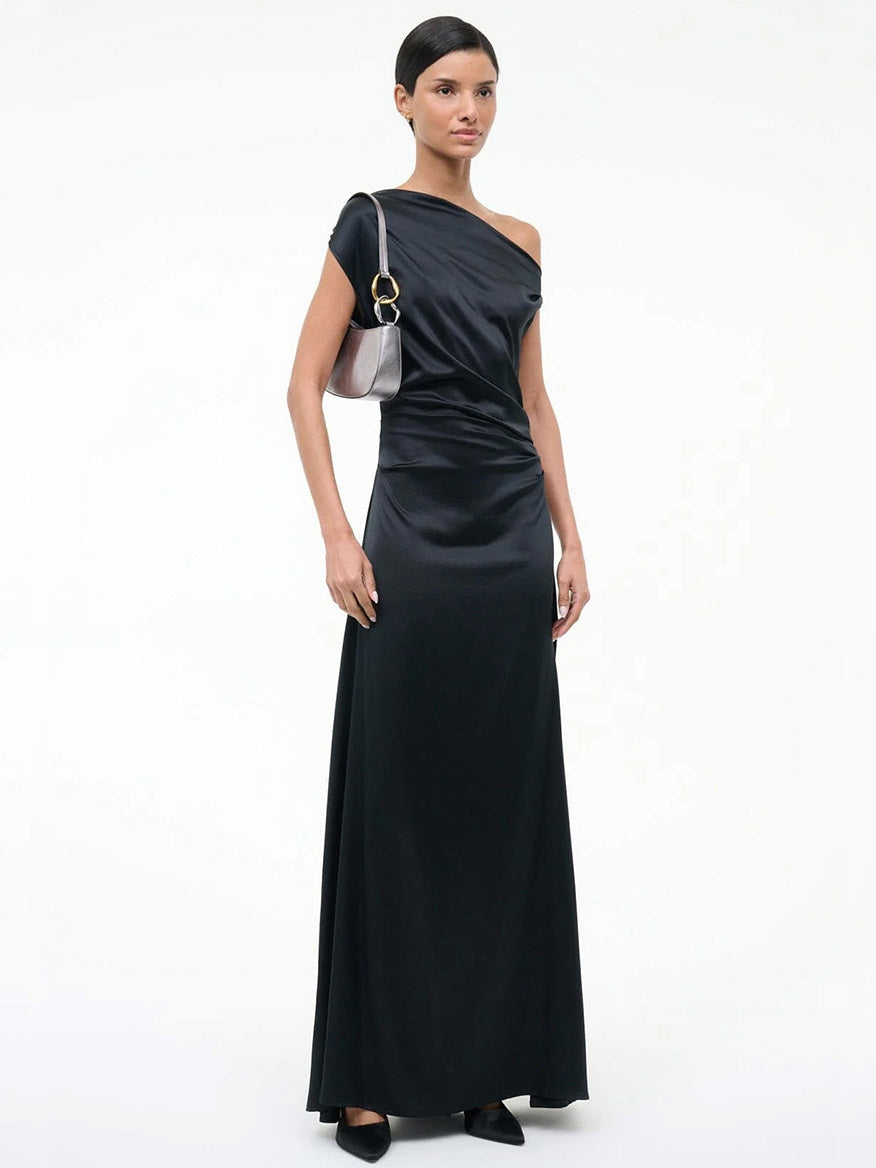 Woman wearing a long black one-shoulder satin gown with black shoes, holding the STAUD Ollie Bag in Pewter with two-tone hardware, standing against a plain white background.