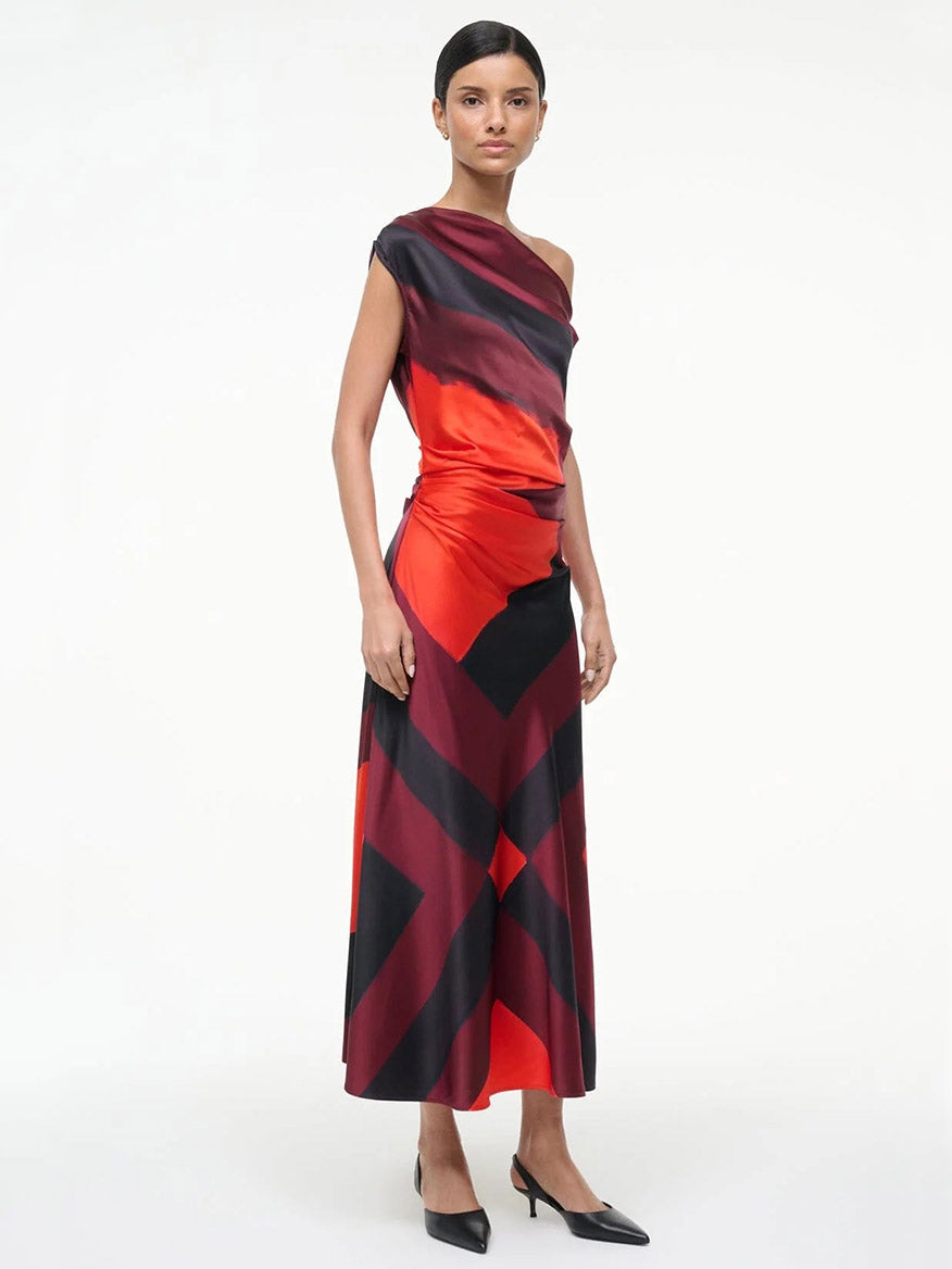 A woman stands in the STAUD Phare Silk Dress in Ruby Prism, an ankle-length, one-shoulder stretch silk dress featuring bold red and black geometric patterns, paired with black heels against a plain white background.
