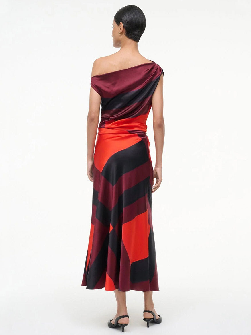 A woman stands facing away, wearing the STAUD Phare Silk Dress in Ruby Prism and black sandals against a plain white background.