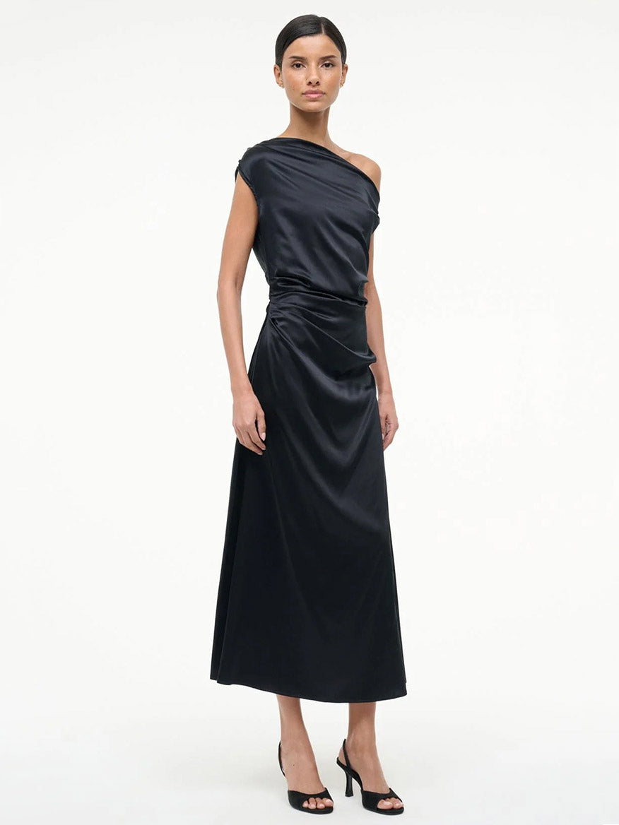 A woman stands against a plain background wearing the STAUD Phare Silk Dress in Black, a one-shoulder, ankle-length stretch silk dress with ruched detailing, paired with black open-toe heels.
