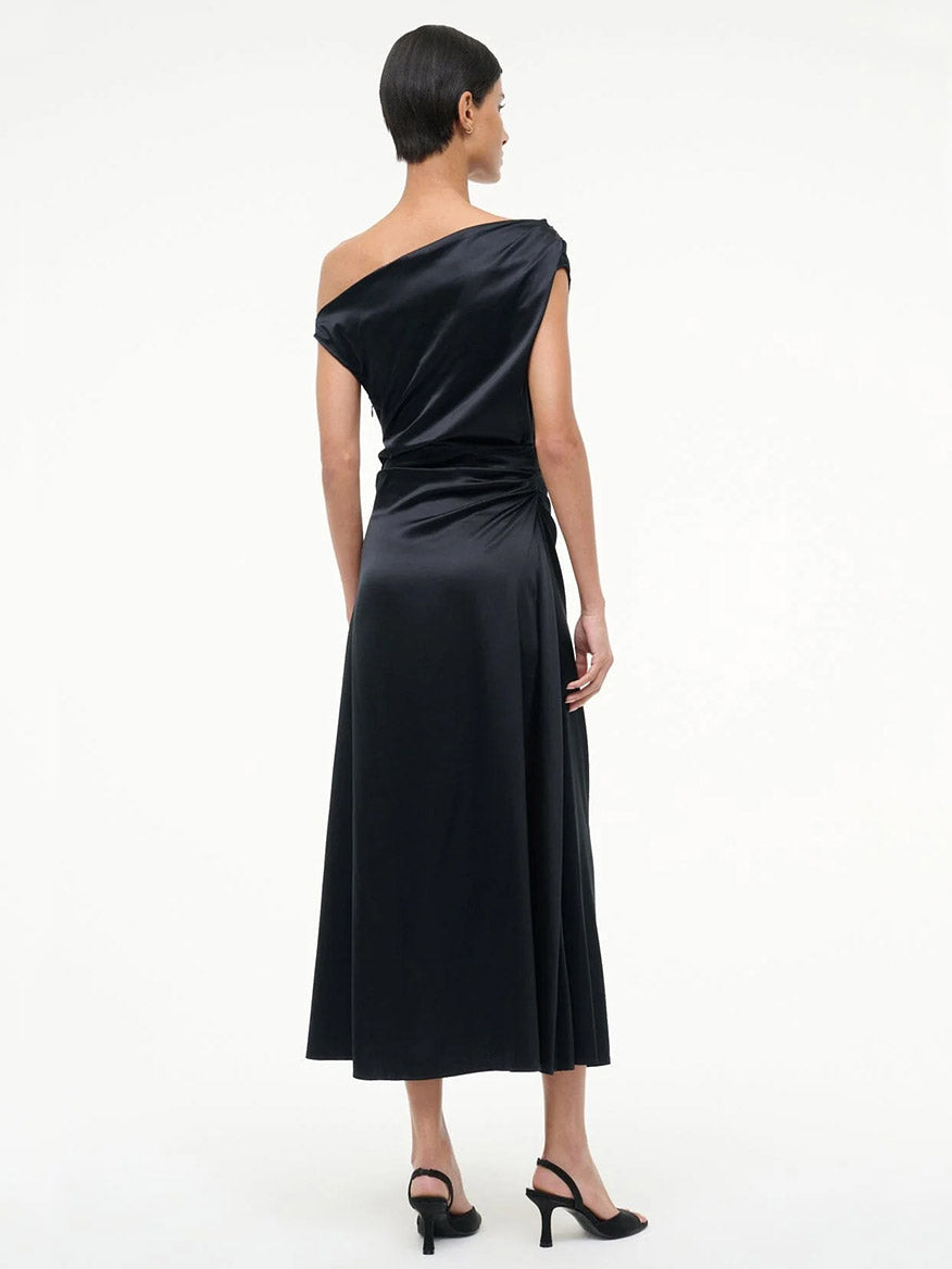 A person with short hair faces away, wearing the STAUD Phare Silk Dress in Black and black heeled sandals against a plain white background.