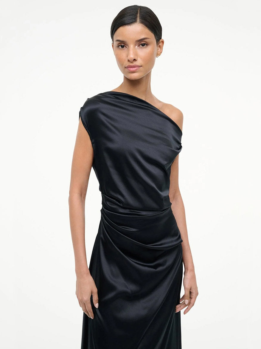 A woman stands against a plain background wearing the STAUD Phare Silk Dress in Black—a sleek, off-the-shoulder design with draped waist detailing perfect for special occasions.