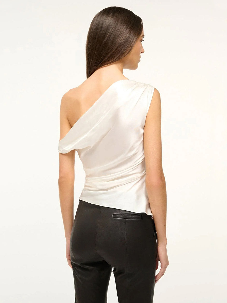 A woman faces away, wearing the STAUD Phare Silk Top in Ivory—a white, drapey one-shoulder silk satin top—and black pants against a plain light background.