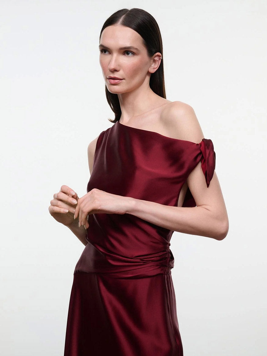 A person wears a STAUD Portia Silk Dress in Carmino, featuring a dark red, one-shoulder design with adjustable shoulder ties.