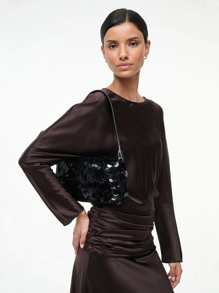 A woman in a dark brown satin long-sleeve dress holds the STAUD Timmy Shoulder Bag in Black with a detachable strap over her shoulder, standing against a plain white background.