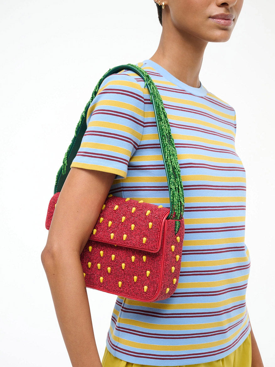 A person in a striped short-sleeve shirt holds the STAUD Tommy Beaded Bag in Strawberry Dream, a playful beaded shoulder bag with green straps and yellow dots that looks like a strawberry—the perfect accessory for any outfit.